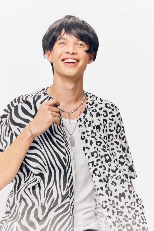 Smiling man wearing a unique zebra and leopard print shirt with layered necklaces, expressing happiness and style against a plain white background.