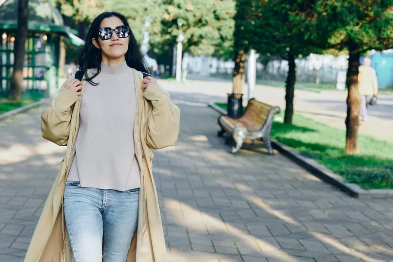 Fashionable street style with a long beige coat, knit sweater, distressed jeans and sunglasses as a confident figure strolls through a sunlit park avenue today