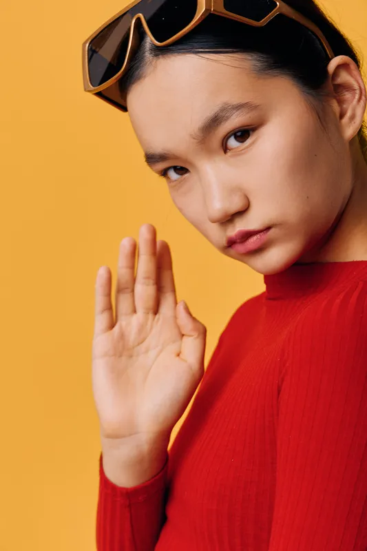Portrait of young woman with sunglasses on head, standing against yellow background, wearing red top, posing with hand raised and serious facial expression in studio lighting.