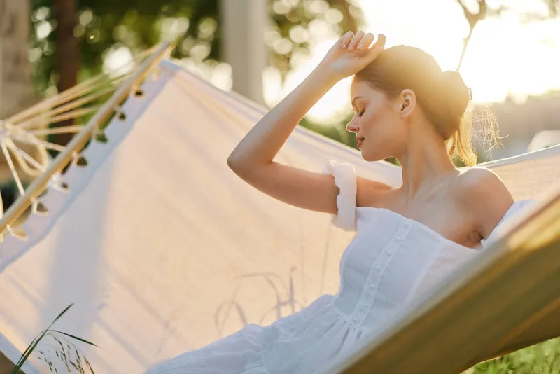 Serene young woman relaxing in a white dress on a hammock, representing summer vibes and tranquility, surrounded by natural greenery and soft sunlight