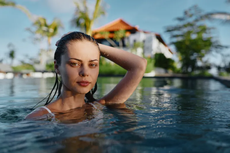 Bright outdoor pool portrait of a woman partially submerged in sunlit water, showcasing a relaxed mood and tranquil summer vibe by a resort setting with palm trees