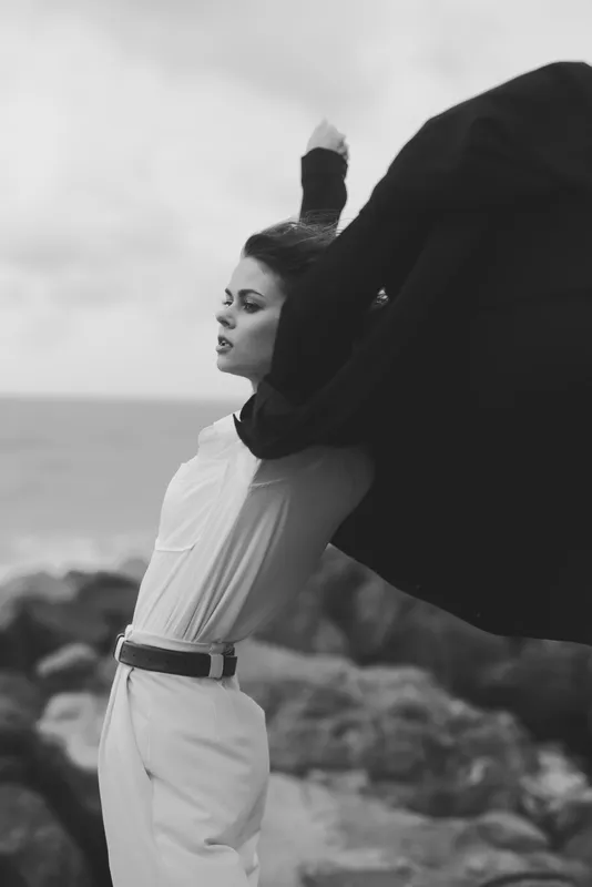 Monochrome fashion shot of a woman in a white dress as a dramatic cape billows, ocean rocks in the background, expressive motion and windwear