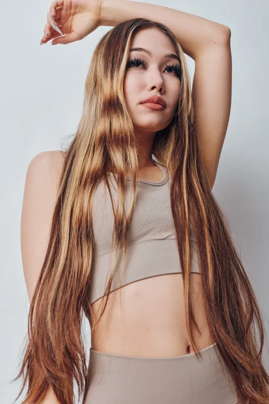 Young woman with long light brown hair poses with raised arm wearing beige fitness wear against plain background for beauty, health, and fashion concepts.