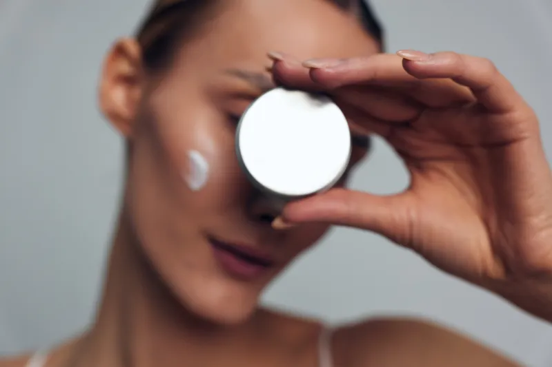 Young woman skincare beauty concept, applying cream with hand on face, holding round cosmetic pad near eye, soft focus portrait, calm clean skin, care routine