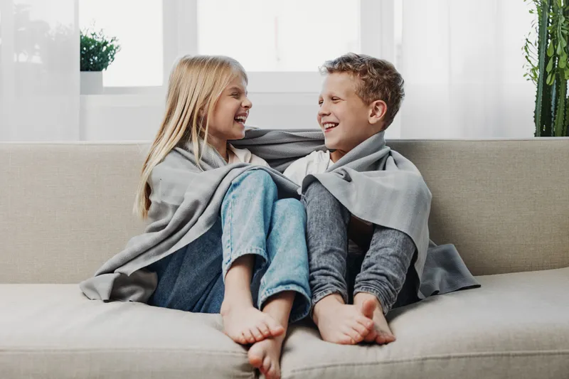 Happy children wrapped in cozy blankets sit barefoot on a sofa, laughing and enjoying a warm moment in a bright living room. Concept of childhood joy and comfort.