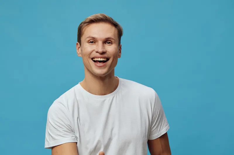 Smiling young man with cheerful expression, wearing casual white t-shirt, posing against plain blue background, studio portrait, happiness, positive emotions, lifestyle concept