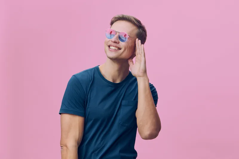 Smiling woman with short hair wearing stylish sunglasses and a casual blue t-shirt, posing against vibrant pink background, cheerful expression, lifestyle portrait, youthful energy, summer fashion.