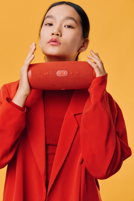 Young woman in red outfit holding portable wireless speaker, posing against orange background with serious expression and modern fashion style, closeup portrait for technology.