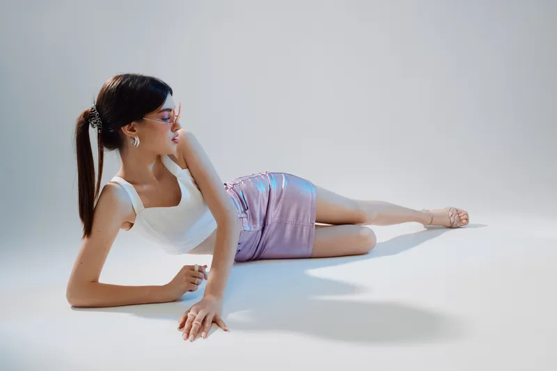 Young woman in a white top and shiny skirt poses lying on the floor against a light background. Concept of beauty, fashion, style, and modern elegance in studio lighting.