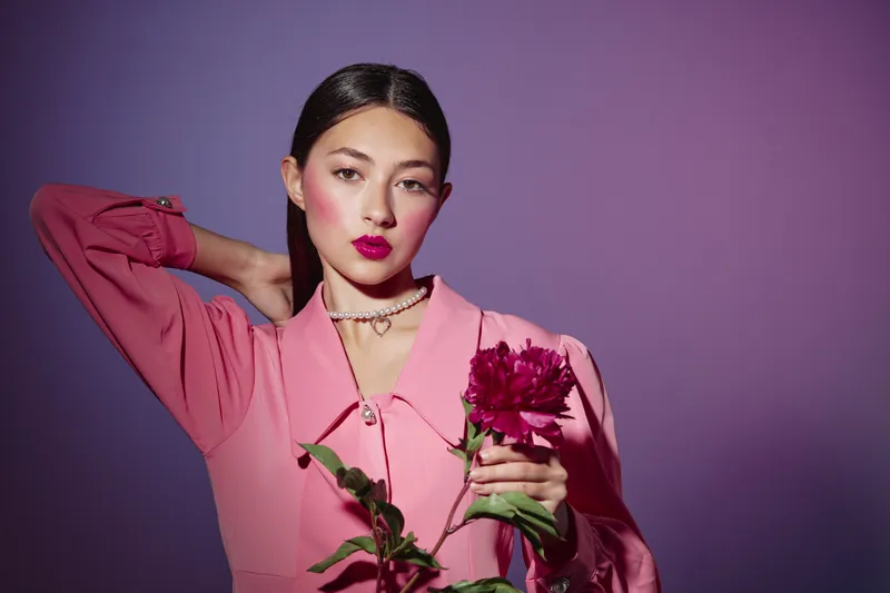 Young woman in pink dress with deep red lipstick poses holding a flower against violet background. Fashion and beauty creative work with intense styling.