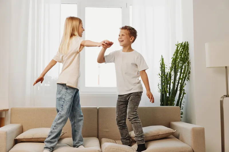 Happy children playing on sofa in bright living room, holding hands and smiling. Boy and girl enjoy carefree moment indoors. Concept of childhood fun, joy, and sibling bonding at home.