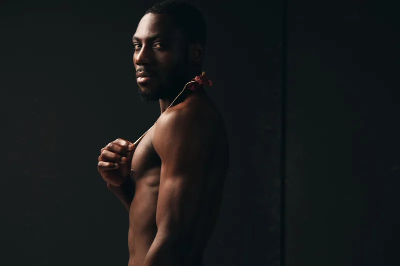 African man with dark skin and muscular body posing shirtless in creative conceptual visual style with serious expression on black background for artistic portrait photography.