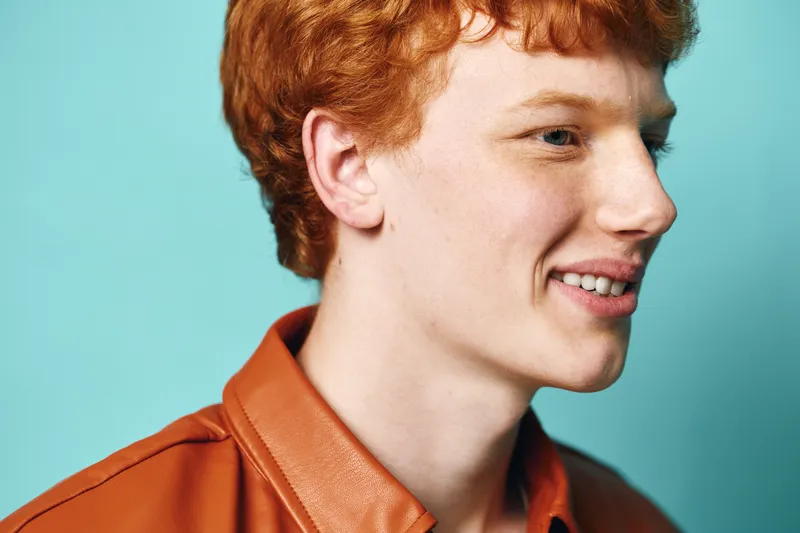 Young man with red hair and freckles wearing brown leather jacket smiling and looking sideways against blue background in closeup portrait for fashion and lifestyle.