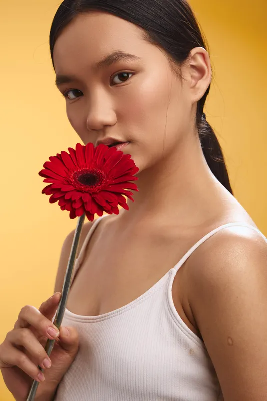 asian woman poses with a red flower held near her face against a warm yellow background, exuding calm elegance, soft expression, and natural beauty.