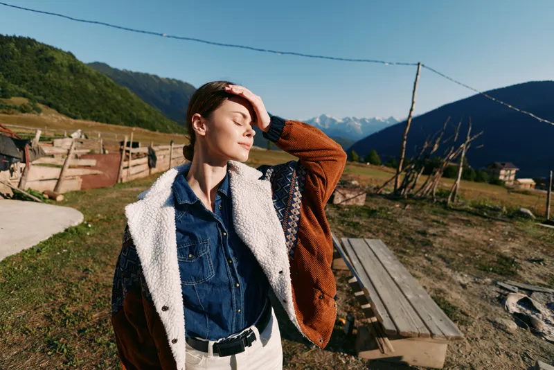 Woman portrait in countryside mountains with sunlight on face, relaxed outdoor fashion look wearing cozy jacket and denim shirt near wooden bench and rustic fence in scenic nature.