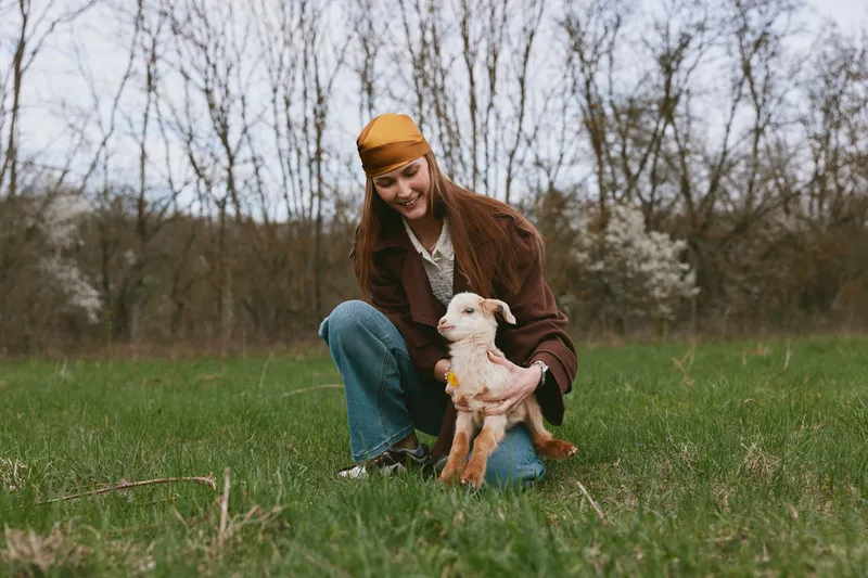 Young woman in casual clothes crouches on green grass while holding a small white goat in a natural outdoor setting during early spring.