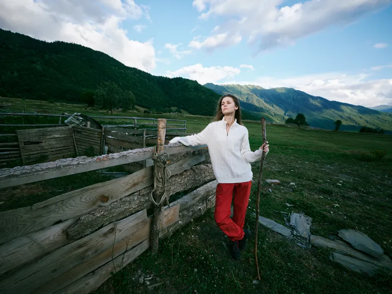 Female traveler leaning on a rustic wooden fence in a green valley with distant mountains, wearing white sweater and red pants, holding a walking stick outdoors