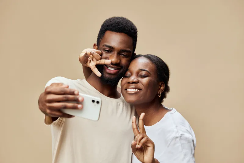 Happy young couple taking a selfie together against a warm beige background, showcasing love and joy in their casual outfits