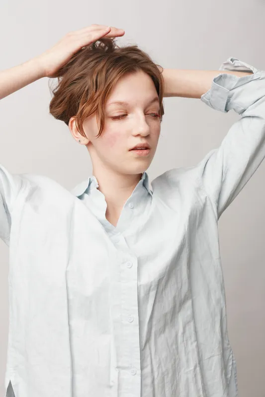 Young woman with short hair in white shirt poses with eyes closed and hands on head. Studio portrait on light background. Concept of relaxation, calmness, and natural beauty.