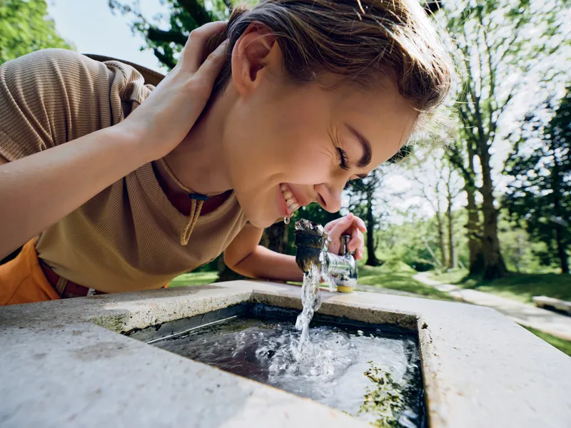 Woman drinking from a stone fountain in a sunlit park, closeup of smiling face and flowing water. Outdoor leisure and hydration captured with natural daylight and greenery.