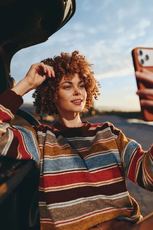 Woman lifestyle selfie curly hair striped sweater sunset outdoor phone held near face as friend adjusts hair in scenic outdoor background soft evening light relaxed smile today