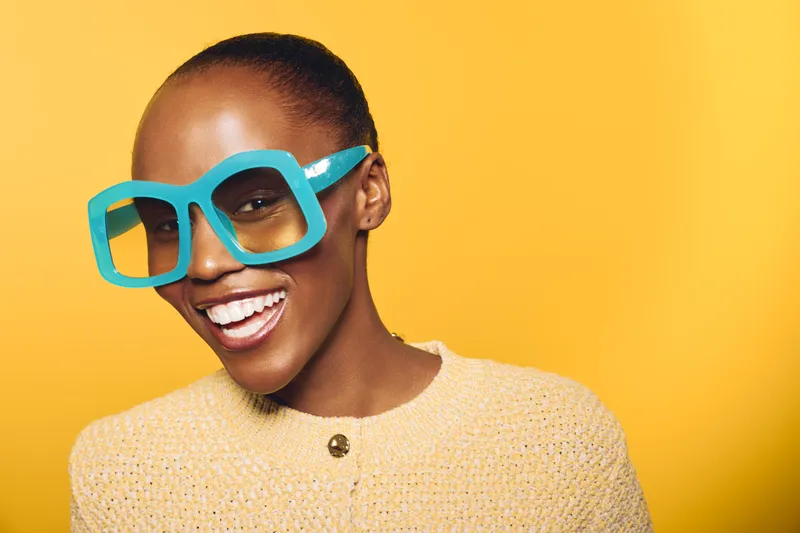 Smiling dark skinned African woman wears large blue glasses and yellow knit sweater. Close-up portrait on yellow background. Concept of fashion, joy, and confident female model with bright style.