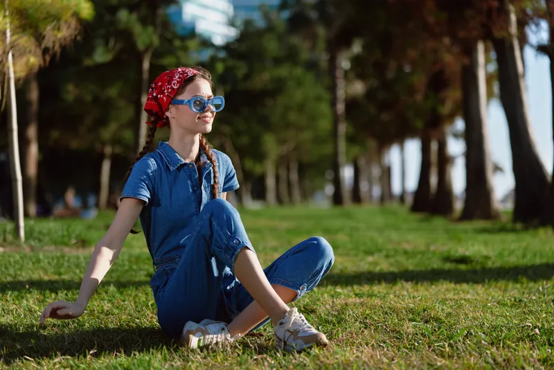 young woman sitting on green grass in park wearing blue jumpsuit and red headscarf enjoying sunny day outdoors with sunglasses in casual relaxed pose