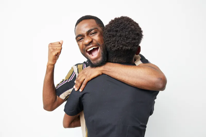 Two black men celebrate joyfully in an embrace, showcasing their friendship and happiness against a clean, white background, capturing a moment of pure excitement.