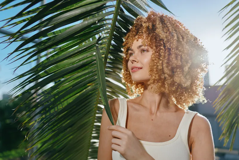 young woman with curly hair standing near palm leaves under bright sunlight, wearing a white tank top, enjoying a sunny summer day outdoors