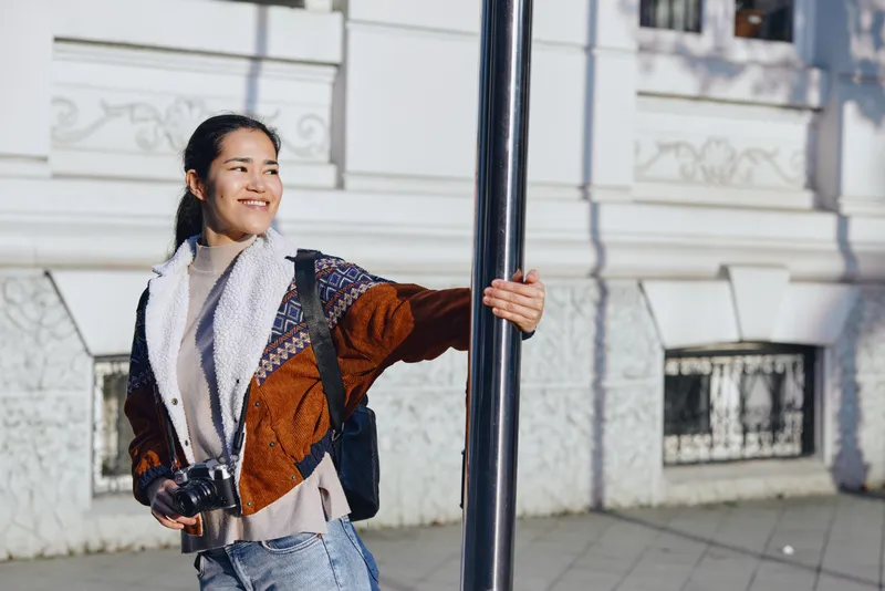 Woman with camera and backpack leaning on a pole on a sunny street, smiling, urban backdrop, casual jacket and scarf, candid outdoor photography capturing travel and daily life
