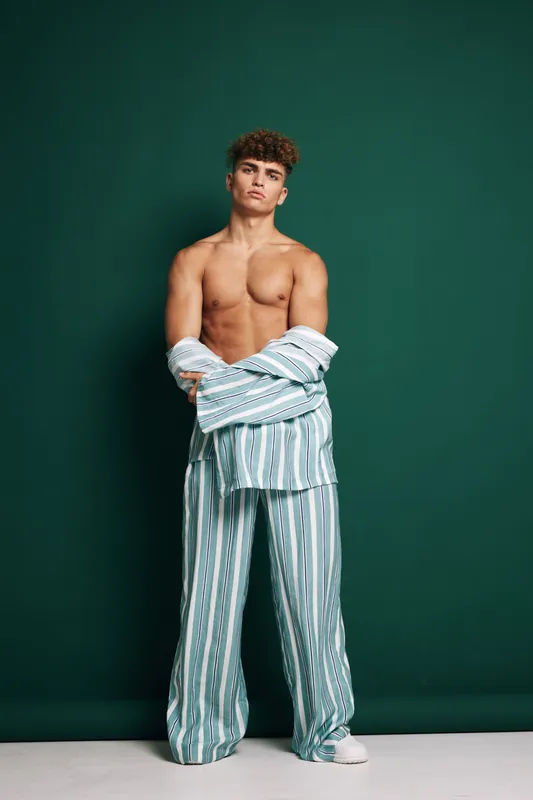 Young man with curly hair posing shirtless wearing striped pajama pants and open shirt against green background. Casual style, confident expression and relaxed posture in studio.