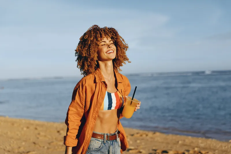 Bright beach scene with a smiling woman in casual orange shirt, crop top and jeans, holding a drink with a straw, sunlit sea breeze, relaxed pose, warm natural colors