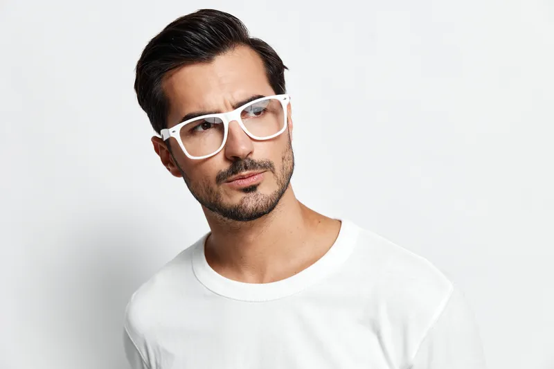 Thoughtful handsome man wearing white t-shirt and stylish glasses looking at camera with serious expression against simple solid light background. Fashion concept.