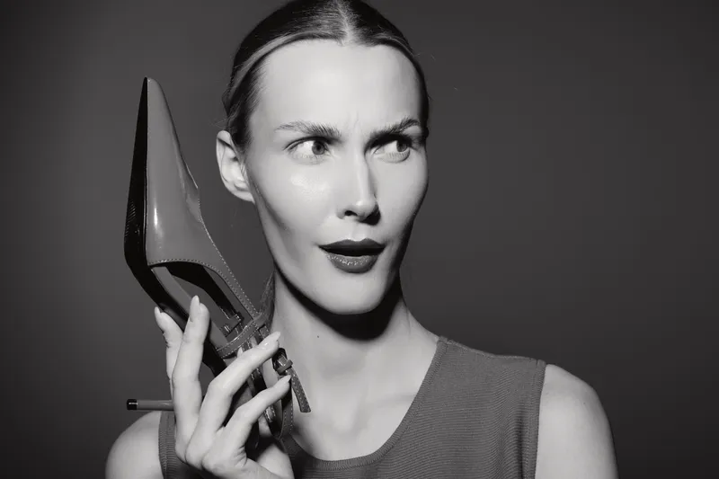 Caucasian white woman with pale skin holds high heel shoe near face with confused expression, studio portrait on dark background.