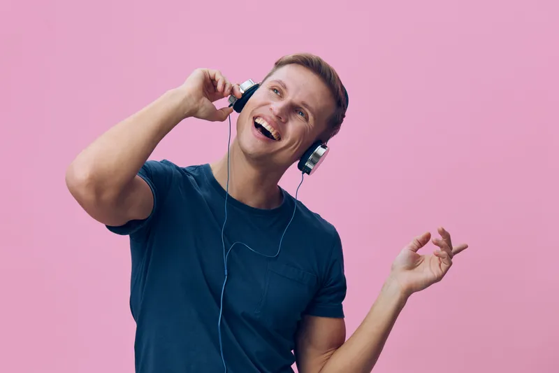 Happy young man wearing headphones enjoying music, smiling broadly, wearing casual blue t-shirt, energetic and cheerful mood, isolated on pink background, studio portrait. People lifestyle concept