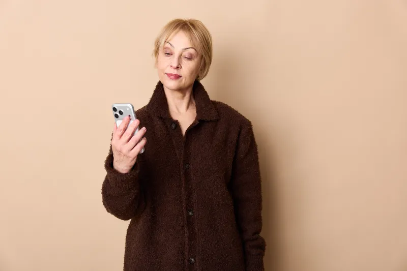 Smiling woman in a dark coat holds a smartphone for mobile technology and communication, in a studio portrait against a beige background, looking at the screen.