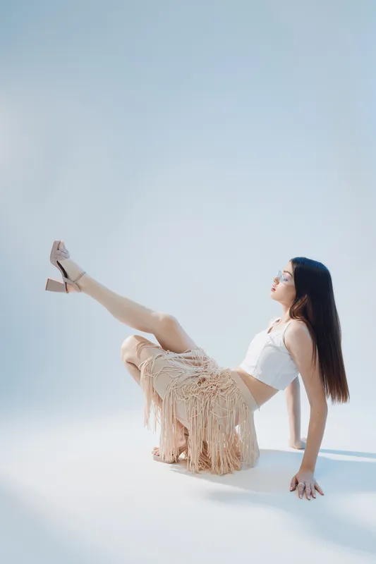 Young woman in white top and beige skirt poses with one leg raised on a light background. Concept of beauty, fashion, and modern style with soft natural lighting.
