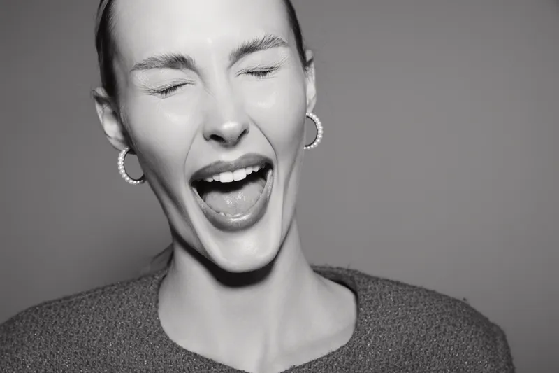 Young white woman with earrings closes eyes and laughs with open mouth against grey background. Concept of joy and happiness.