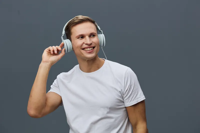 Smiling young man wearing white t-shirt and headphones listen to music, looking away against plain gray background, casual pose, cheerful expression, lifestyle, relaxed mood, portrait. People concept