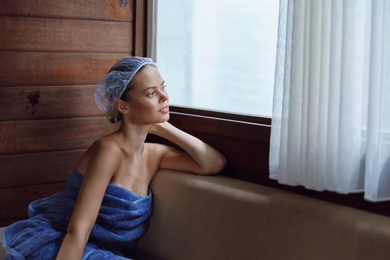 relaxed woman wrapped in blue towel gazes through window with soft natural light in cozy wooden interior, calm contemplative mood at home during daytime.