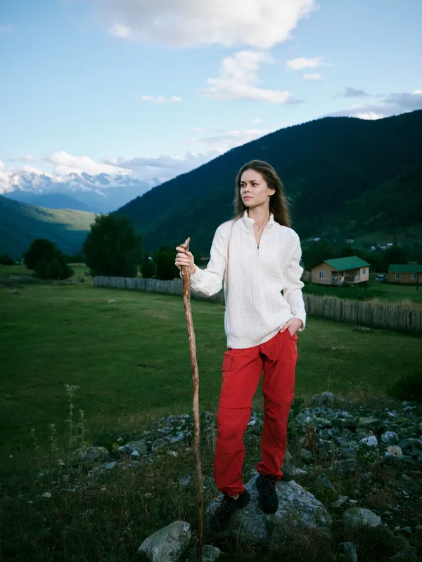 Woman standing on rocky hill with walking stick in a green valley, mountains in distance, white sweater and red pants, exploring nature and open countryside
