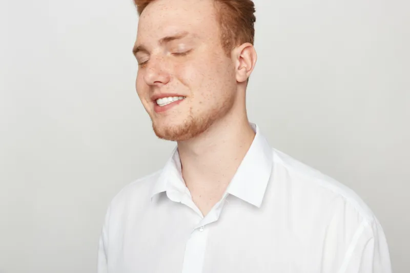 Young man with red hair and beard smiles with closed eyes wearing white shirt on light background. Concept of happiness, relaxation, and positive emotions in casual style.