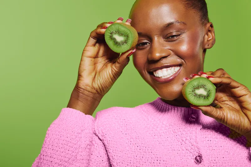 Smiling dark skinned African American woman in pink sweater holds two halves of kiwi fruit near face on green background. Concept of healthy lifestyle, nutrition, and fresh organic food.