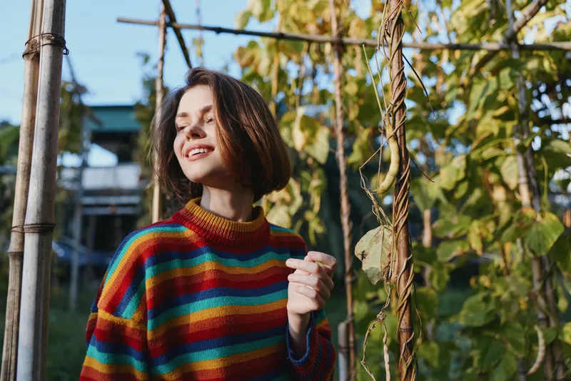 Woman smiling in a garden wearing a colorful striped sweater and casual outfit, young adult with bob haircut leaning by a vine trellis in warm autumn sunlight for lifestyle portrait.