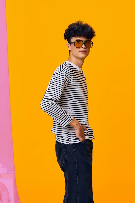 Young man wearing striped shirt and sunglasses stands confidently against bright orange background, showcasing casual style and relaxed attitude with curly hair.