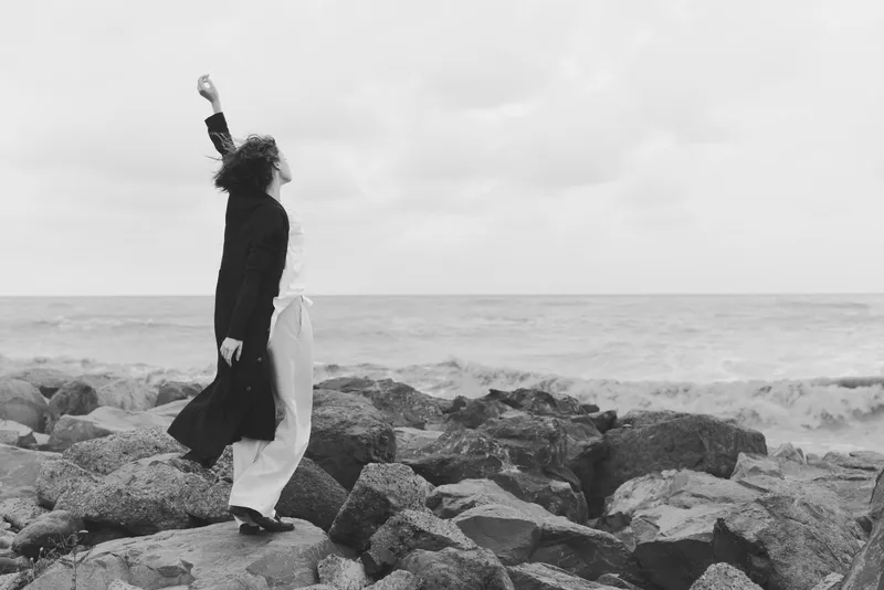 woman on rocks by the sea, dress blowing in wind, stance facing horizon, black and white outdoor scene conveying mood, solitude, resilience, and cinematic atmosphere of coastal wind and vast water