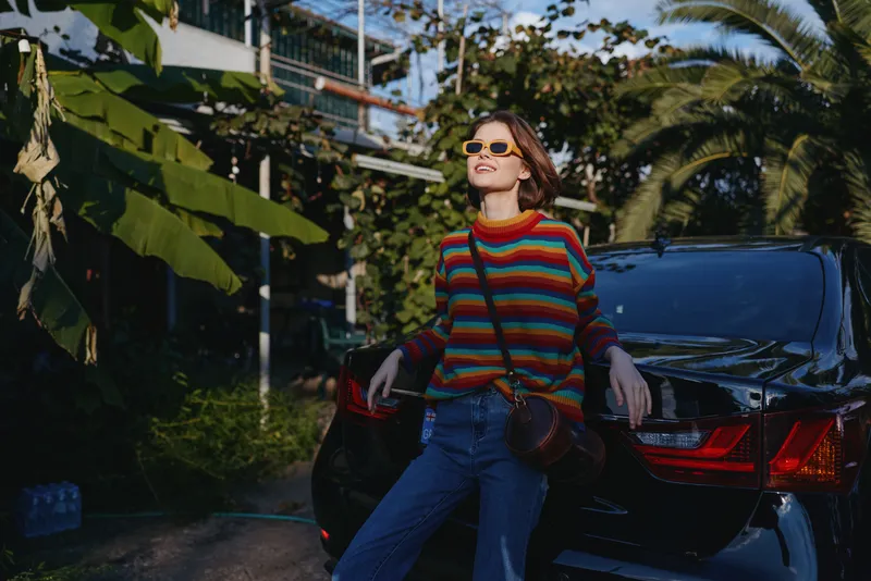Woman in a colorful striped sweater and sunglasses leaning against a black car in a tropical outdoor city setting, smiling in a casual outfit with crossbody bag for travel lifestyle vibe.