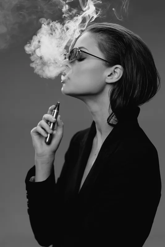 Black and white portrait of a woman vaping with smoke around her face and sunglasses, expressing calm and modern style in monochrome tones.