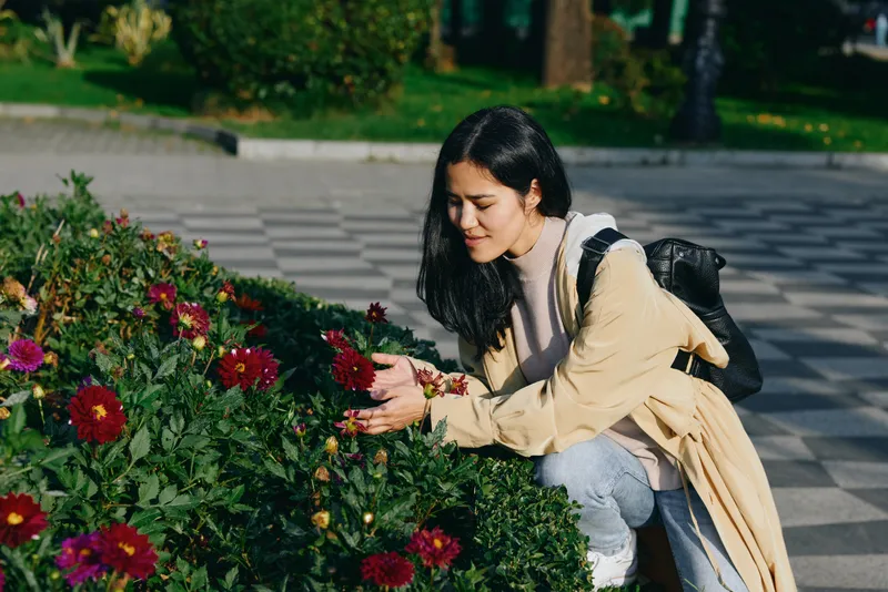 Woman kneels beside blooming flowers in a park, touching petals with curiosity, capturing a serene moment of nature, calm exploration and outdoor gardening.