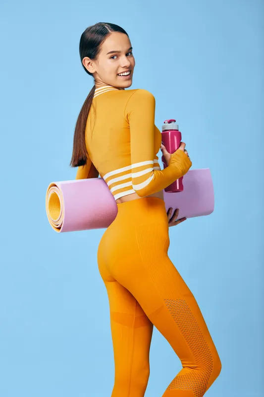 Woman fitness portrait in bright orange activewear holding a pink yoga mat and shaker bottle, smiling over her shoulder in studio lighting, sport concept emphasizing strength and posture.